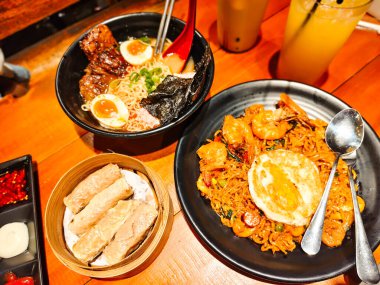 A hearty bowl of delicious fried seafood ramen noodles and beef ramen in tori paitan soup with a side dish of shrimp dim sum on a bamboo tray. Asian cuisine at a restaurant