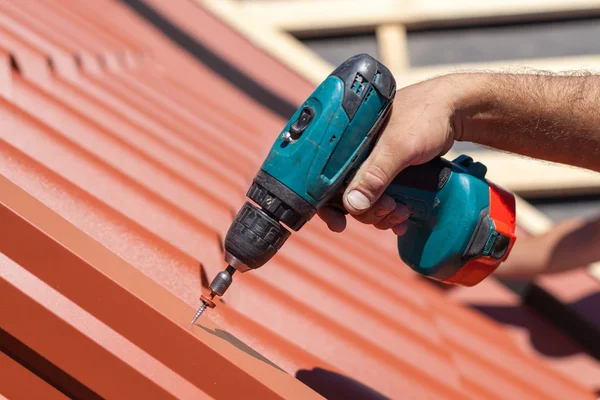 Operaio su un tetto con trapano elettrico l'installazione di piastrelle metalliche rosse sulla casa di legno — Foto Stock