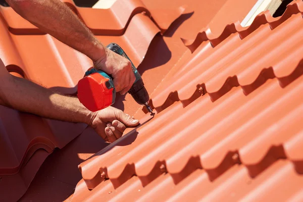 Trabalhador em um telhado com broca elétrica instalando telha de metal vermelho na casa de madeira — Fotografia de Stock