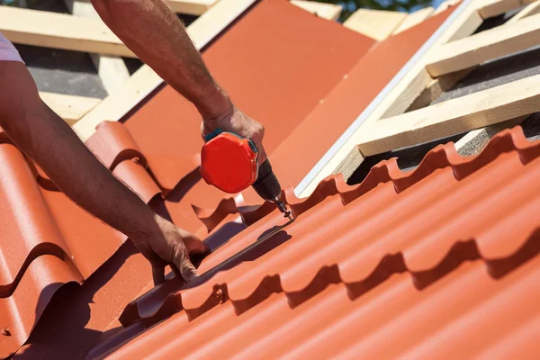 Trabalhador em um telhado com broca elétrica instalando telha de metal vermelho na casa de madeira — Fotografia de Stock