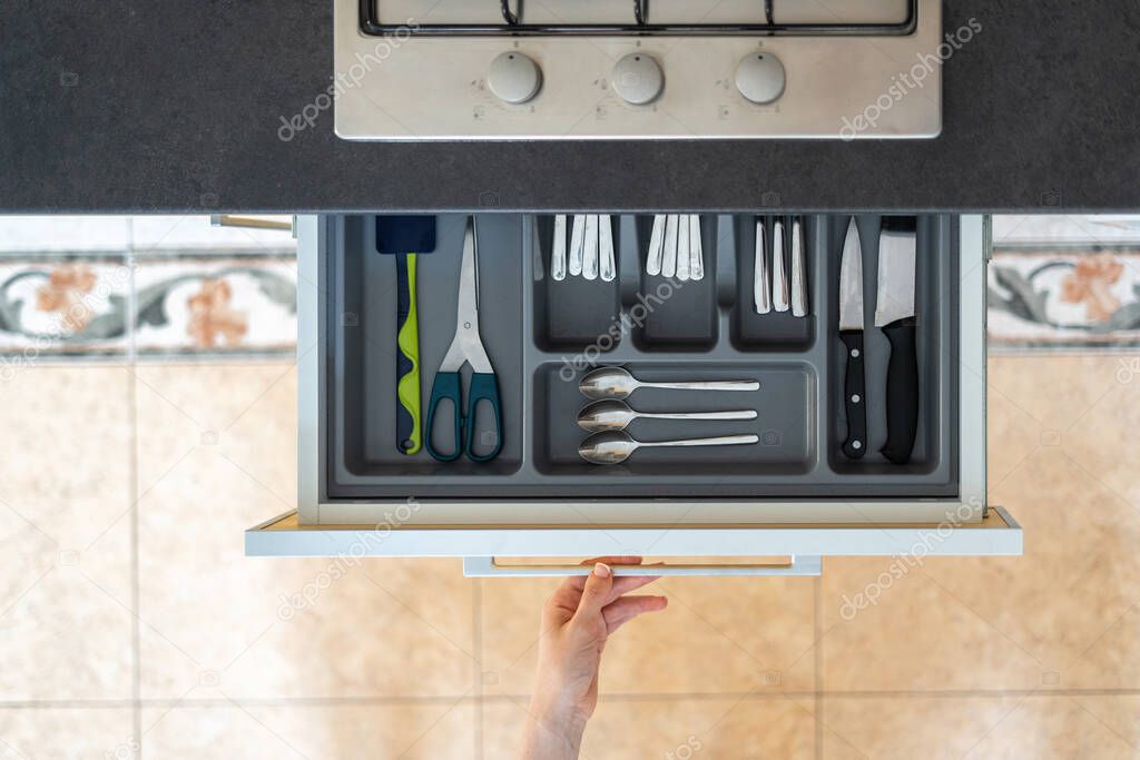 Top view of woman hand opened cutlery drawer with different spoons, knives and forks under countertop with gas stove cooker on modern kitchen