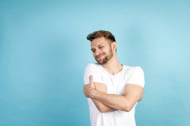 Kişisel destek, ilgi ve sevgi kavramı. Beyaz tişörtlü mutlu genç adam mavi fotokopi arkaplanda tek başına duruyor, kendine sarılıyor, kapalı gözlerle rüya gibi bir yüz yapıyor.