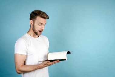 Başarılı sınav konsepti. Beyaz tişörtlü genç adam şok olmuş bir yüz ifadesi takınıyor, ödev defterine bakıyor, mavi fotokopi çekilen arka planda tek başına duruyor.