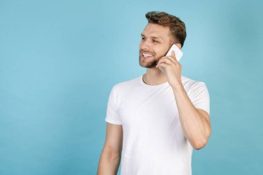 Beyaz tişörtlü, gülümseyen bir yetişkinin modern akıllı telefondan konuşmasının ve mavi fotokopi arkaplanında tek başına durmasının yan görüntüsü. İletişim konsepti