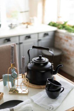 Sıcak bir içecek ve siyah bardakların yanında duran metal çaydanlığın dikey görüntüsü ve sıcacık mutfaktaki masada, bulanık dolapların karşısında duran ev dekorasyonu.