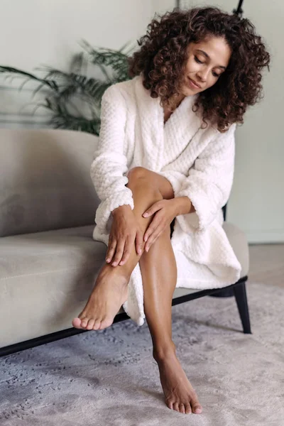 Skincare and body care concept. Vertical view of calm african american woman in bathrobe sitting at home, touching soft skin on legs after shaving and waxing hair removal procedure, smiling nice