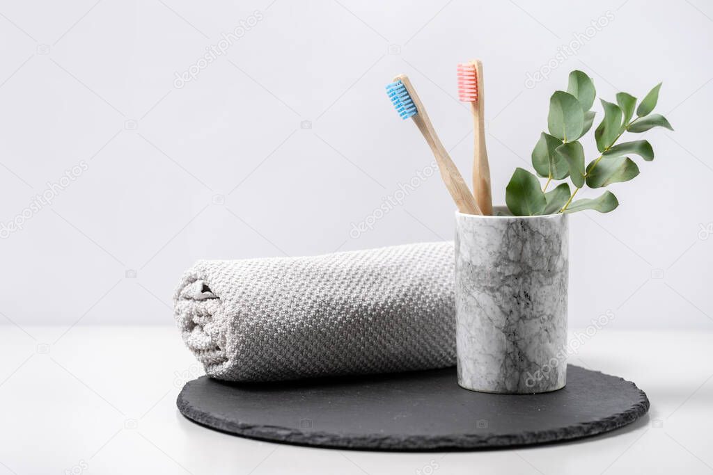 Concept of biodegradable objects. Two bamboo toothbrush in cup near bathroom towel and eucalyptus plant on black plate stand against white copy space background. Oral and dental care