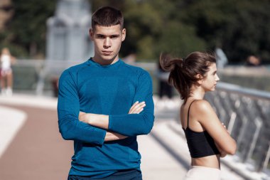 Ciddi bir genç atletizmin portresi, kamera karşısında el pençe divan duran, bir kadınla birlikte açık havada yapılan spor sonrası bulanık şehir eğitimine karşı duran.