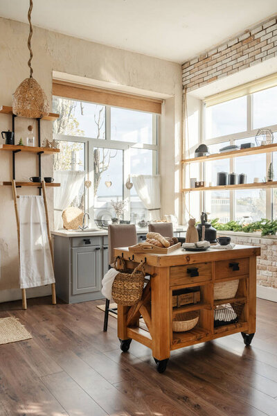Vertical side view of stylish sunny kitchen interior with kitchenware, home decor and accessories, wooden table with homemade pastry food against cupboards and windows
