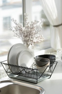 Vertical view of plates, clean dishes, kitchenware utensil on metal dryer rack against blurred bouquet in white vase in cozy home at modern kitchen