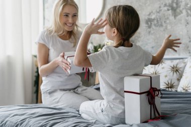 Back view of little daughter hiding present behind her back, looking at happy mother with gift box in hands and sitting on comfort bed. Concept of celebrating holidays at home