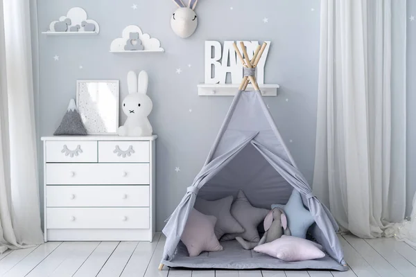 Happy childhood concept. Kids playroom with pillows in tent, white wooden commode, bunny toys and house decor in apartment with scandinavian interior design