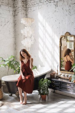 Concept of morning routine. Vertical view smiling young woman sitting on classic bath at comfortable bathroom with green houseplants in flower pots, decor at commode and interior design in boho style clipart