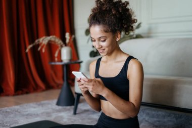 Mutlu ve gülümseyen genç Afro-Amerikan kadınlarının yan görünüşü, modern akıllı telefonlarda online uygulama kullanması, hafta sonları evdeki oturma odasında oturması, internet üzerinden arkadaşları ve ailesiyle iletişim kurması.