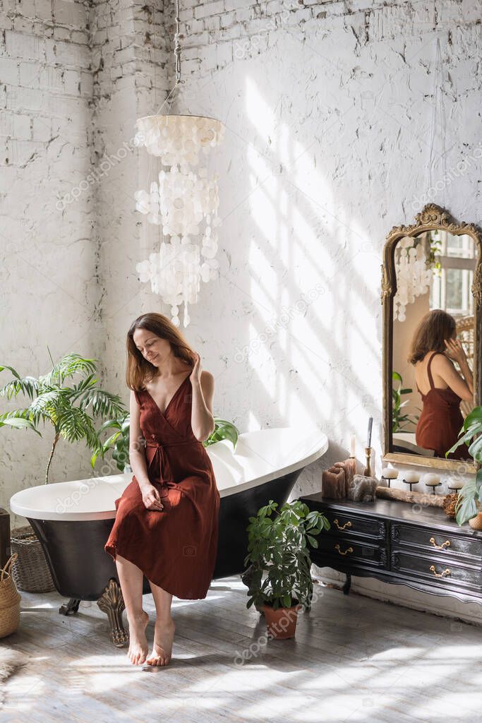 Concept of morning routine. Vertical view smiling young woman sitting on classic bath at comfortable bathroom with green houseplants in flower pots, decor at commode and interior design in boho style
