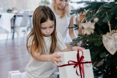 Geleneksel kış tatili kavramı. Sakin küçük kız hediye kutusunu elinde tutarken, Noel ağacının yanında otururken annesi gülümsüyor, odasını dekore ediyor ve yeni yıla hazırlanıyor.