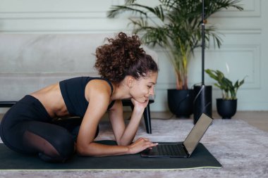 Çevrimiçi spor eğitimi konsepti. Düşünceli yetişkin Afro-Amerikan kadın modern dizüstü bilgisayarda yoga egzersizi izleyip evde antrenman yapmaya hazırlanıyor.