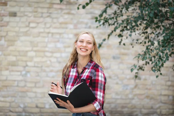 Happy university student girl standing near campus building with copybooks and smiling wide. Charming young woman looking at camera walking outdoors after courses, classes, faculty or lecture