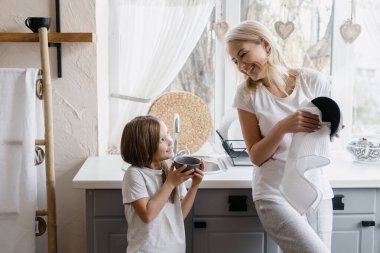 Ev işlerine yardım ederken mutlu bir anne bulaşıkları yıkıyor ve gülümseyen kızına bakıyor. Anne tabakları siliyor ve küçük kızla evde vakit geçiriyor. Mutfak dolaplarının yanında duruyor.