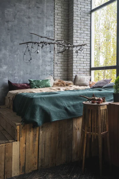 Vertical view of comfort bed with soft pillows and plaid near beverage on coffee table under large window. Bedroom with loft interior, light bulb garland on wooden branch, home decor in cozy apartment