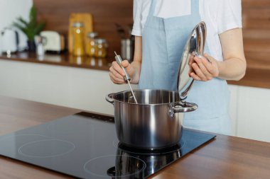 Önlüklü bir kadının tencerede lezzetli yemekler pişirmesini cam seramik yüzeyi olan elektrikli soba kullanmasını ve modern mutfakta bulanık fotokopi alanının arka planına karşı durmasını.