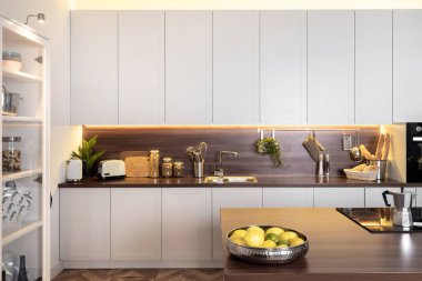 Ahşap tezgahın üzerinde beyaz mutfak dolapları, lavabonun yanında yiyecek malzemeleri, kahve makineli elektrikli soba ve masada taze limon.