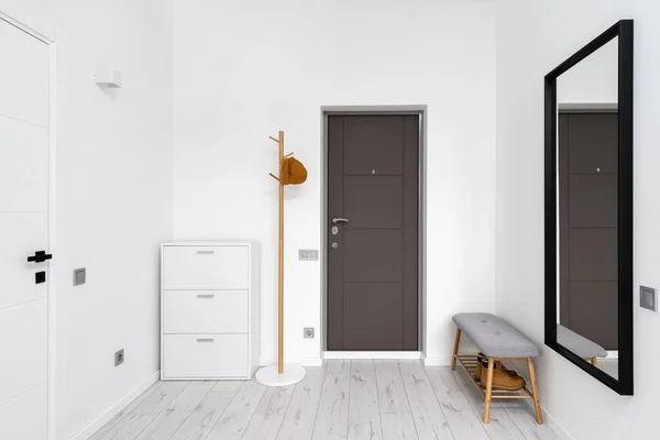 Minimalist hallway with modern interior design in contemporary apartment. Comfortable shoe storage bench near black frame mirror on white wall. Wooden coat hanger in hall with chest drawers