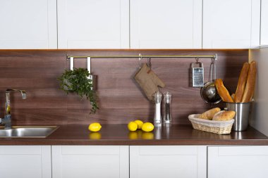 Beyaz mutfak dolapları, ahşap tezgah, metalik lavabo, mutfak gereçleri, limonlar ve sepette taze ev yapımı ekmek.