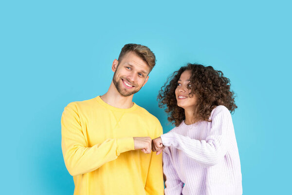 Happy afro american woman bump fists with handsome european man standing on blue background with copy space. Two friends make greeting gesture and smiling wide. Friendship concept