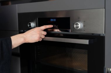 Modern mutfakta duran, fırın aletlerini kullanan, kontrol panelindeki programı değiştiren, ev yapımı yemek pişirirken sergilenecek zamanı seçen kadın görüntüsü.