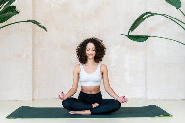 Spor kıyafetleri giyen, yoga stüdyosundaki egzersiz minderinde meditasyon pozu veren sakin, Afro-Amerikan bir kadın. Sportif kadın huzurlu ve sakin bir yüz yapıyor. Yogi asana çalışıyor.