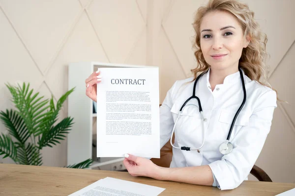 Woman doctor with stethoscope holding contract, showing document on camera. Healthcare check up and insurance concept. Female medic sitting in clinic office behind desk, wearing in white coat uniform