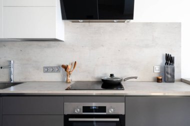 Cihazlı mutfak, elektrikli fırın kaputu, fırın ve lavabo. Ahşap kaşık ve bıçaklara yakın mermer tezgahta, cam seramik yüzey üzerinde tava, beyaz dolapların altında.