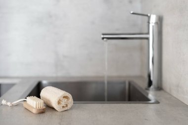 Geri dönüşüm, azaltma, konsepti yeniden kullanma. Çevre dostu bulaşık süngeri ve modern mutfaktaki mermer tezgahta plastiksiz küçük ahşap fırça, bulanık arkaplanda açık deniz musluğuna karşı.