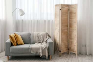 Modern iç tasarımı olan rahat bir oturma odası, rahat bir kanepe, yumuşak ekose, sarı yastıklar, ahşap paravan ve pencereye karşı tüllü beyaz bir lamba.