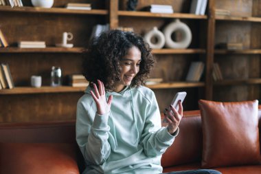 Video çağrısı konsepti. Mutlu Afro-Amerikalı kadın arkadaşlarıyla çevrimiçi sohbet ediyor, modern akıllı telefon kullanıyor, kameraya selam veriyor. Evdeki kanepede oturan gülümseyen genç kız.