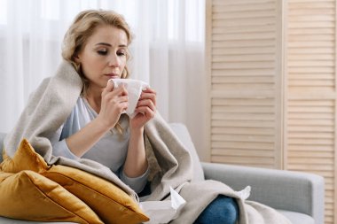 Evde hastalık kavramı. Sağlıksız ve üzgün bir kadın battaniyeyle kaplı bir şekilde kanepeye oturur ve elinde peçeteyle gezer. Üzgün kadının ateşi ve solunum yolu enfeksiyonu var.