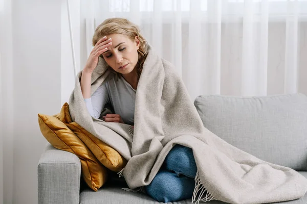 Concept of sickness and disease. Tired, ill woman with headache touch forehead, checking temperature, covered with blanket. Female having migraine, fever and flu symptoms. Lady sit on sofa at home