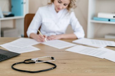 Sağlık kontrollerini hatırlatma kavramı. Steteskoplu çalışma masasına odaklan. Kadın doktor kişisel kabinede oturuyor, günlüğe yazıyor, tıbbi geçmişi dolduruyor ya da hasta hafızasını kaybediyor.