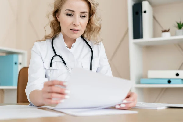 Paperwork and medical insurance concept. Selective focus on concentrated woman doctor in white medical uniform with patient treatment plan. GP working behind desk in hospital office