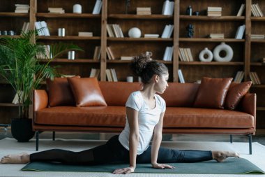 Afro-Amerikan kadın spor kıyafetlerini giymiş, bacaklarını esnetip oturma odasında yerde otururken bacaklarını ayıran profilden bir görüntü. Günlük antrenmandaki kadın, evdeki spor minderinde yoga eğitimi alıyor.
