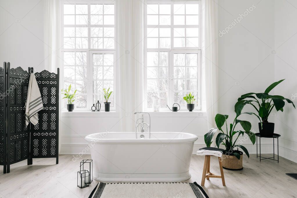 Home decor ideas in contemporary bathroom design. Empty freestanding bathtub against large windows, wooden folding screen and green house plants on floor. Concept of classic bath in modern apartment