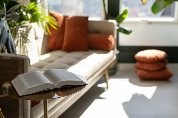 Open book lying on minimalist coffee table in living room against blurred background with comfortable sofa, orange pillows and green potted plants. Concept of cozy apartment in boho style
