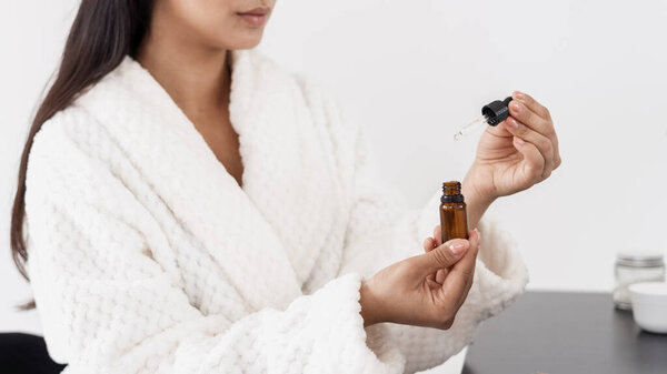 Концепция красоты. Обрезанный снимок молодой женщины в халате с бутылкой эфирного масла для сияющей и увлажненной кожи. Лечение лица гиалуроновой кислотой, коллагеном и витамином Е, панорамный вид