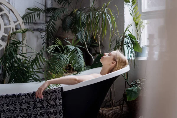 美丽的概念 轻松自在的年轻女子在舒适舒适的豪华公寓里洗澡 梦幻般的女性躺在浴缸里 一上午呆在浴室里 把温室里的植物放进罐子里 — 图库照片