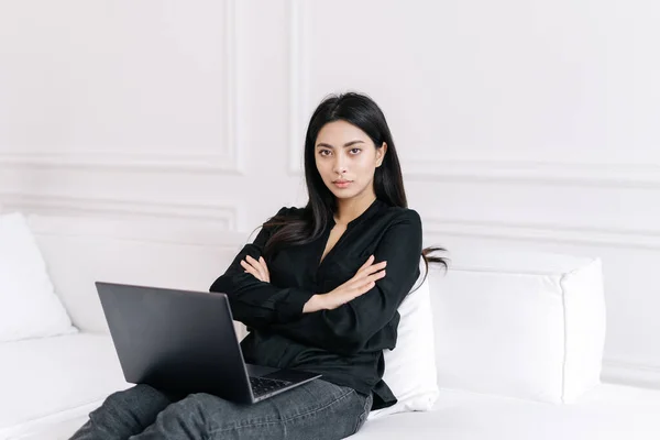 Confident young asian business woman working remotely on laptop at home, sitting on couch with arms crossed and looking directly at camera with confident expression. Female freelancer sitting on sofa