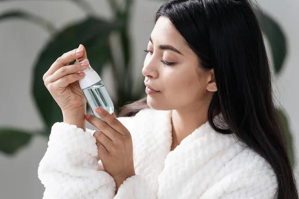 Concept of natural beauty and skincare procedure. Side portrait view of young asian woman looking at bottle with organic skin care cosmetic. Female in white bathrobe spending morning in bathroom