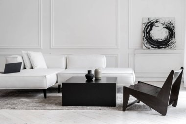 Üzerinde açık dizüstü bilgisayar olan şık beyaz bir kanepe, çağdaş sandalye ve masa bulunan minimalist bir odada, siyah ve beyaz soyut resimlerle süslenmiş bir daire ve masada iki vazo.