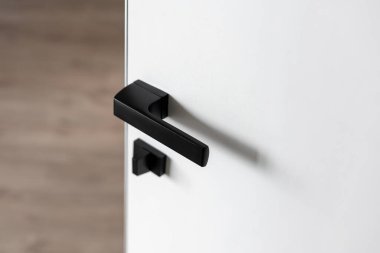 Kırpılmış beyaz görüntü, minimalist siyah mat kapı kolu ve bulanık ahşap zemin zeminli güvenlik kilidiyle oda kapısını açtı. Ev güvenliği ve modern apartman tasarımı kavramı