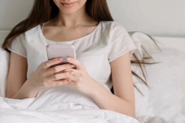 インターネット中毒の概念 現代のスマートフォンでオンラインアプリを使用して笑顔の若い女性の作物のビュー 女性でベッドの上に座っているライトベッドルーム 過ごす朝で居心地の良い家 — ストック写真